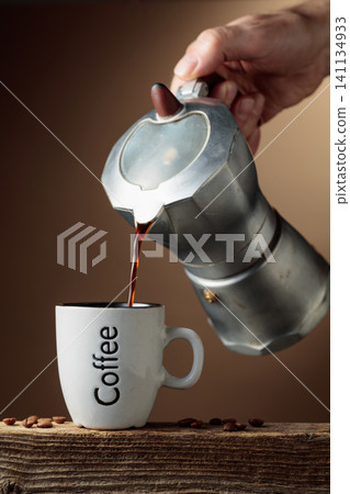 Black coffee is poured from a classic Italian aluminium moka pot into a coffee cup. Black coffee is poured from a classic Italian aluminium moka pot into a coffee cup. 141134933