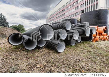 Construction workers are preparing materials at a site where black and orange pipes are lined up on the ground near a building on a cloudy day Construction workers are preparing materials at a site where black and orange pipes are lined up on the ground near a building on a cloudy day 141134977