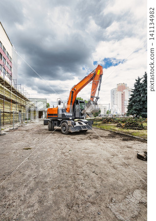 A construction vehicle digs into the ground at a building site while workers prepare the area. Tall buildings and clouds surround the site. Excavator at work A construction vehicle digs into the ground at a building site while workers prepare the area. Tall buildings and clouds surround the site. Excavator at work 141134982