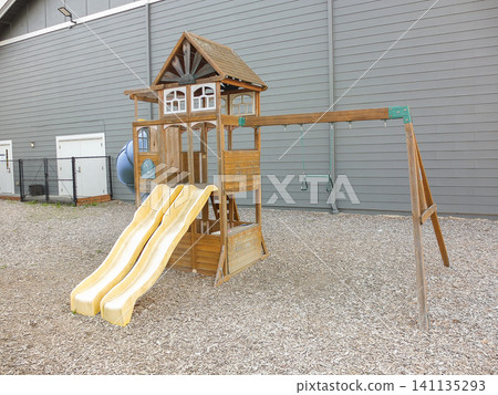 Wooden children playground set with swings and climbing wall on wood chip ground by a gray wall - June 6, 2026 Milton WA, USA Wooden children playground set with swings and climbing wall on wood chip ground by a gray wall - June 6, 2026 Milton WA, USA 141135293
