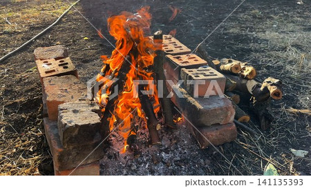 Small campfire with wooden sticks brightly burn at sunny summer day. Gentle flames and glowing embers create warm vibrant atmosphere. The scene conveys outdoor adventure and elemental beauty of fire Small campfire with wooden sticks brightly burn at sunny summer day. Gentle flames and glowing embers create warm vibrant atmosphere. The scene conveys outdoor adventure and elemental beauty of fire 141135393