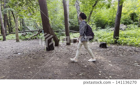 Young girl going along path at summer forest. Female hiker with backpack walking among trail at wild nature. The scene reflecting adventure, travel and connection with wild nature. Slow motion 141136270
