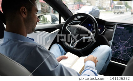 Male businessperson reading book during riding on electrical vehicle with autopilot at rainy day. Successful businessman improving knowledge while ride an autonomous self driving electric car at city Male businessperson reading book during riding on electrical vehicle with autopilot at rainy day. Successful businessman improving knowledge while ride an autonomous self driving electric car at city 141136282