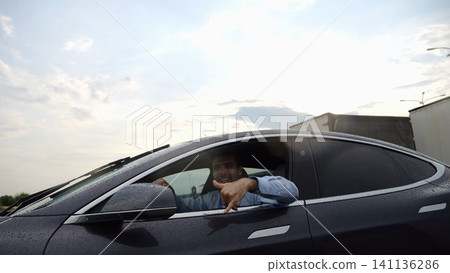 Young businessman driving on electric car in circular path on asphalt road after rain. Happy manager showing grimaces and fooling around while riding on modern auto. Concept of green innovation Young businessman driving on electric car in circular path on asphalt road after rain. Happy manager showing grimaces and fooling around while riding on modern auto. Concept of green innovation 141136286