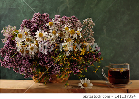 Still life with wild medicinal herbs on a wooden table in a basket and cups of infusion or tea, a bouquet of St. John's wort, oregano, mint, and chamomile, a collection of useful herbs Still life with wild medicinal herbs on a wooden table in a basket and cups of infusion or tea, a bouquet of St. John's wort, oregano, mint, and chamomile, a collection of useful herbs 141137354