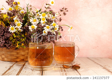 Still life with wild medicinal herbs on a wooden table in a basket and cups of infusion or tea, a bouquet of St. John's wort, oregano, mint, and chamomile, a collection of useful herbs Still life with wild medicinal herbs on a wooden table in a basket and cups of infusion or tea, a bouquet of St. John's wort, oregano, mint, and chamomile, a collection of useful herbs 141137377