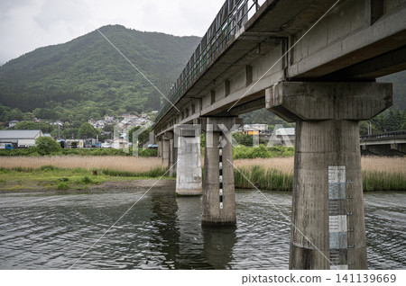 Under a cloudy, rainy sky, the scenery includes a bridge pier with a water level gauge, a concrete bridge crossing the river, and the mountain range visible in the distance. 141139669