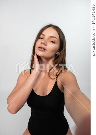 Young woman taking selfie, posing in black bodysuit, showing confidence and natural beauty with glowing skin 141141489