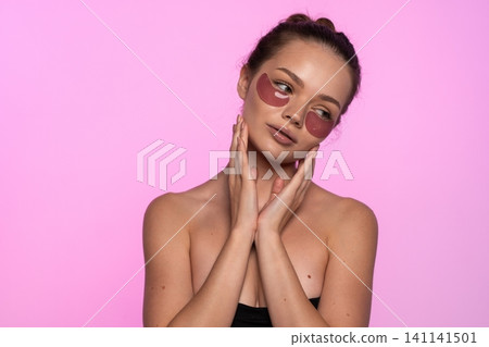 Young woman applying pink eye patches for skincare and self-care on a vibrant pink background Young woman applying pink eye patches for skincare and self-care on a vibrant pink background 141141501