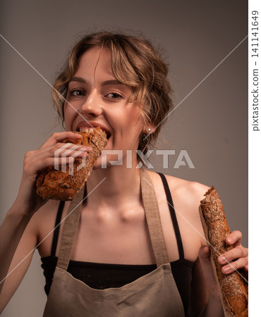Hungry young woman in apron biting fresh crusty sourdough baguette, enjoying a delicious snack Hungry young woman in apron biting fresh crusty sourdough baguette, enjoying a delicious snack 141141649