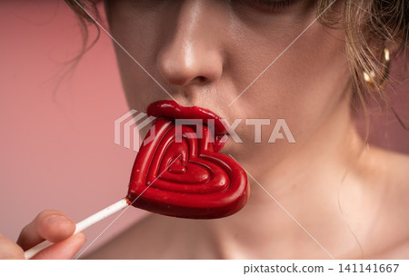 Woman's seductive red lips playfully holding a heart-shaped lollipop, symbolizing romance, desire, and sweet love Woman's seductive red lips playfully holding a heart-shaped lollipop, symbolizing romance, desire, and sweet love 141141667