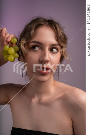 Young woman holding green grapes, looking away, with modern jewelry, posing on a purple background Young woman holding green grapes, looking away, with modern jewelry, posing on a purple background 141141668