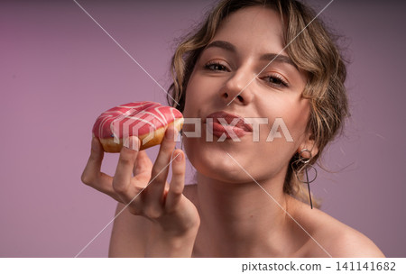 Woman holding a brightly colored pink frosted donut, anticipating pleasure from eating the sweet treat Woman holding a brightly colored pink frosted donut, anticipating pleasure from eating the sweet treat 141141682