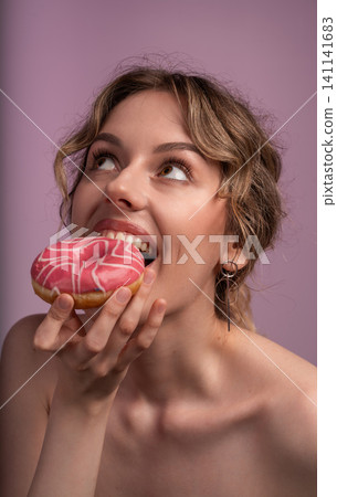 Woman taking a bite of a pink glazed donut, smiling and looking up. Expressing joy and craving for sweets Woman taking a bite of a pink glazed donut, smiling and looking up. Expressing joy and craving for sweets 141141683