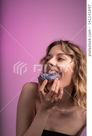 Woman joyfully biting into a purple glazed donut, showing enjoyment and indulgence with sweet food Woman joyfully biting into a purple glazed donut, showing enjoyment and indulgence with sweet food 141141697