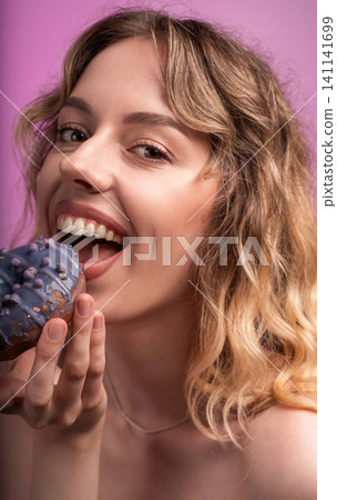 Happy young woman enjoying a sweet purple doughnut, having fun eating dessert with a cheerful expression Happy young woman enjoying a sweet purple doughnut, having fun eating dessert with a cheerful expression 141141699