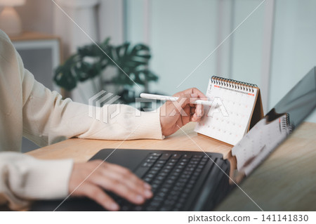 Worker planning to write paper work report presentation meeting dates on calendar, using laptop computer at wooden desk in office, organizing timeline for corporate business project Worker planning to write paper work report presentation meeting dates on calendar, using laptop computer at wooden desk in office, organizing timeline for corporate business project 141141830