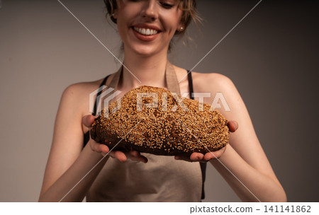 Young woman baker holding and presenting a freshly baked dark artisan bread with sesame seeds, smiling Young woman baker holding and presenting a freshly baked dark artisan bread with sesame seeds, smiling 141141862