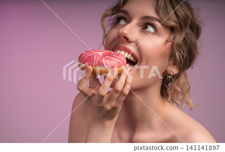Young woman eating a pink glazed donut, indulging in a sweet dessert, craving more sugary treats and enjoying a snack Young woman eating a pink glazed donut, indulging in a sweet dessert, craving more sugary treats and enjoying a snack 141141897