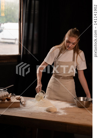 Woman pouring milk into flour on a wooden table, preparing dough for baking bread, cooking in kitchen Woman pouring milk into flour on a wooden table, preparing dough for baking bread, cooking in kitchen 141141902