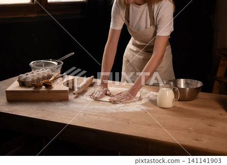 Woman in an apron kneading dough on a wooden table, surrounded by baking ingredients like flour, eggs, and milk Woman in an apron kneading dough on a wooden table, surrounded by baking ingredients like flour, eggs, and milk 141141903