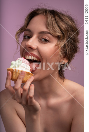 Young woman enjoying sweet dessert, eating a colorful cupcake with gusto and happy expression on pink background Young woman enjoying sweet dessert, eating a colorful cupcake with gusto and happy expression on pink background 141142006