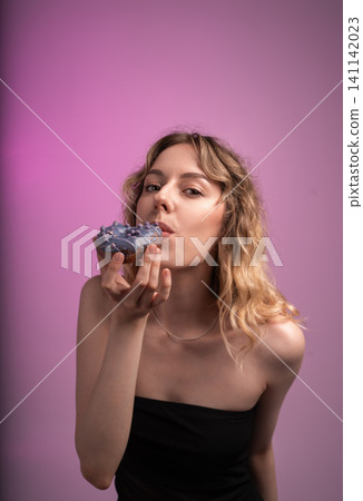 Young woman enjoying sweet donut with purple frosting and sprinkles on a vibrant pink background 141142023
