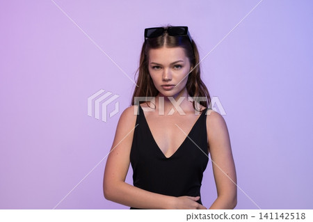 Confident young woman posing in black dress with sunglasses on head against purple background Confident young woman posing in black dress with sunglasses on head against purple background 141142518