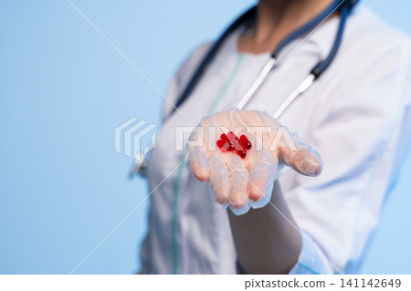 Doctor's gloved hand holding red gel capsules, symbolizing medication, treatment, and medical support in healthcare Doctor's gloved hand holding red gel capsules, symbolizing medication, treatment, and medical support in healthcare 141142649