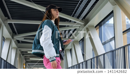 Young woman posing on pedestrian bridge in trendy varsity jacket and pink pants, angled profile with hand on hip and baseball cap accent, confident expression and urban fashion vibe against industrial Young woman posing on pedestrian bridge in trendy varsity jacket and pink pants, angled profile with hand on hip and baseball cap accent, confident expression and urban fashion vibe against industrial 141142682