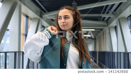 Caucasian woman on steel pedestrian bridge wearing teal varsity jacket and backpack, headphones around neck, thoughtful gaze with subtle smile, modern truss architecture and glass windows, candid Caucasian woman on steel pedestrian bridge wearing teal varsity jacket and backpack, headphones around neck, thoughtful gaze with subtle smile, modern truss architecture and glass windows, candid 141142688