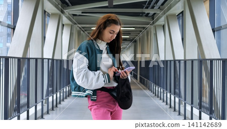 Young woman verifying eticket on smartphone while standing on pedestrian bridge, focused eyes scanning screen and holding small black bag, daylight through glass panels and modern transit context Young woman verifying eticket on smartphone while standing on pedestrian bridge, focused eyes scanning screen and holding small black bag, daylight through glass panels and modern transit context 141142689