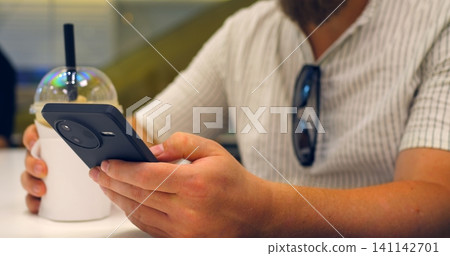 Adult holding smartphone and takeaway drink while seated at cafe table, focused on mobile ordering with black phone, iced beverage cup, striped shirt, and sunglasses hanging from collar. Adult holding smartphone and takeaway drink while seated at cafe table, focused on mobile ordering with black phone, iced beverage cup, striped shirt, and sunglasses hanging from collar. 141142701