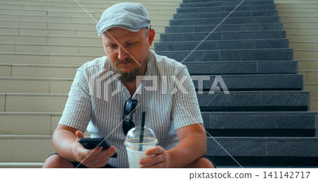 Man sitting on stairs checking smartphone while holding iced drink cup, casual urban portrait with striped shirt, cap, and sunglasses resting on chest in city setting, relaxed everyday moment 141142717