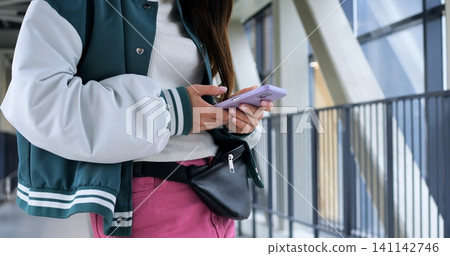 Close midshot of woman checking phone for schedule and notifications, varsity jacket and belt bag visible, focused on screen while leaning on industrial railing in bright corridor. Close midshot of woman checking phone for schedule and notifications, varsity jacket and belt bag visible, focused on screen while leaning on industrial railing in bright corridor. 141142746