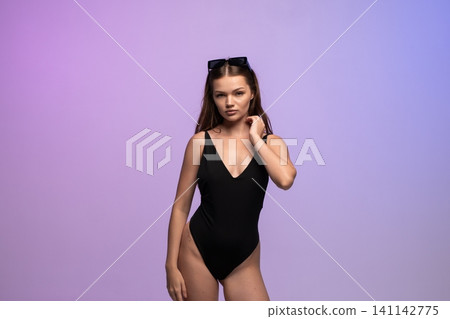 Young woman in black swimsuit with sunglasses posing against purple background, representing summer fashion and beauty Young woman in black swimsuit with sunglasses posing against purple background, representing summer fashion and beauty 141142775