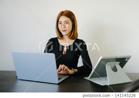 Young professional Asian woman multitasking on laptop and tablet in a modern workspace, showcasing productivity, digital work, and business lifestyle. Young professional Asian woman multitasking on laptop and tablet in a modern workspace, showcasing productivity, digital work, and business lifestyle. 141143226