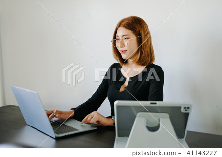 Young professional Asian woman multitasking on laptop and tablet in a modern workspace, showcasing productivity, digital work, and business lifestyle. 141143227
