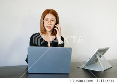 Asian woman on video call using smartphone while working on laptop in modern office, representing remote work, online meeting, and digital communication. Asian woman on video call using smartphone while working on laptop in modern office, representing remote work, online meeting, and digital communication. 141143235