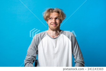 Young man standing against a blue background, expressing confusion and uncertainty with a humorous facial expression, shrugging his shoulders and looking away Young man standing against a blue background, expressing confusion and uncertainty with a humorous facial expression, shrugging his shoulders and looking away 141143383