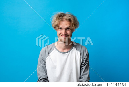 Young man with messy blonde hair and a playful, uncertain expression on a blue background, portraying surprise, humor, or confusion with a quirky smile Young man with messy blonde hair and a playful, uncertain expression on a blue background, portraying surprise, humor, or confusion with a quirky smile 141143384