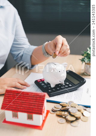 Mortgage budget planning with white piggy bank on calculator, hand adding coin, red house model, contract papers, and coins, ideal for home loan savings, down payment, property finance. Mortgage budget planning with white piggy bank on calculator, hand adding coin, red house model, contract papers, and coins, ideal for home loan savings, down payment, property finance. 141143385
