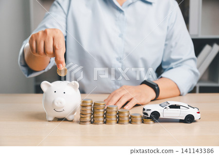 Person putting a coin into a piggy bank beside a white toy car and coin stacks, representing saving money for car purchase, down payment, vehicle finance, and auto loan planning. Person putting a coin into a piggy bank beside a white toy car and coin stacks, representing saving money for car purchase, down payment, vehicle finance, and auto loan planning. 141143386