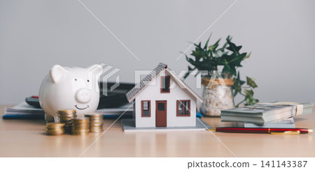 White piggy bank with small house model, coin stacks, cash and pen on office table, representing mortgage savings, home budget, real estate investment, and financial planning. White piggy bank with small house model, coin stacks, cash and pen on office table, representing mortgage savings, home budget, real estate investment, and financial planning. 141143387