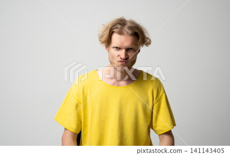Frustrated young man in a vibrant yellow t-shirt looking directly at the viewer, showing annoyance, anger, and a mean expression, dealing with negativity and a bad mood Frustrated young man in a vibrant yellow t-shirt looking directly at the viewer, showing annoyance, anger, and a mean expression, dealing with negativity and a bad mood 141143405