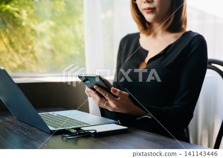 Woman using smartphone while working on laptop at desk with coffee. Business technology, mobile communication, remote work, freelancer lifestyle and modern workspace concept. Woman using smartphone while working on laptop at desk with coffee. Business technology, mobile communication, remote work, freelancer lifestyle and modern workspace concept. 141143446