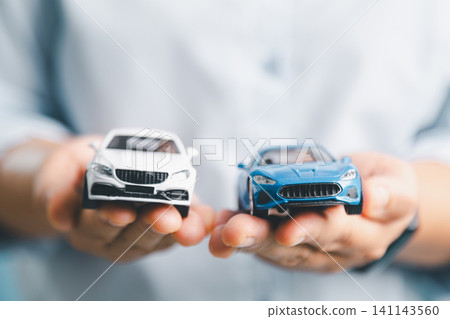Hands holding blue and white toy cars, symbolizing car comparison, auto insurance options, vehicle financing, purchase decision, dealership service, and car loan concept. Hands holding blue and white toy cars, symbolizing car comparison, auto insurance options, vehicle financing, purchase decision, dealership service, and car loan concept. 141143560