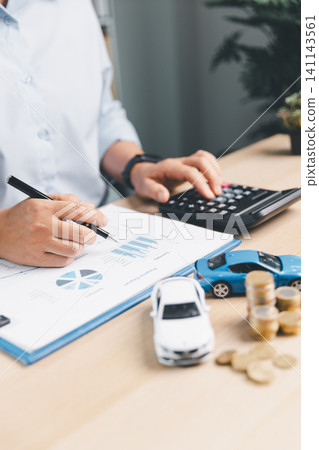 Business person reviewing vehicle finance report with calculator, toy cars, and coin stacks on desk, representing car loan planning, auto insurance, and purchase cost analysis. 141143561