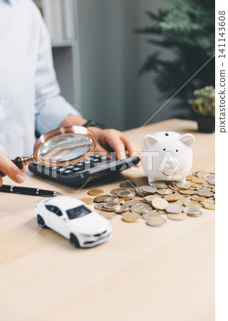 Business person using magnifying glass to review car savings, piggy bank, calculator, coins, and white toy car, representing auto loan analysis and vehicle finance planning. Business person using magnifying glass to review car savings, piggy bank, calculator, coins, and white toy car, representing auto loan analysis and vehicle finance planning. 141143608