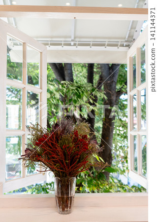 A stylish flower arrangement is displayed in a white, open window overlooking a beautiful garden with fresh greenery. A stylish flower arrangement is displayed in a white, open window overlooking a beautiful garden with fresh greenery. 141144371